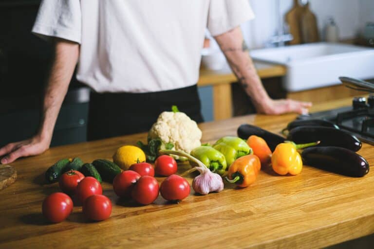 Frisches, vielfältiges Gemüse – Tomaten, Gurken, Paprika, Blumenkohl, Auberginen und Knoblauch – liegt auf einem Holztisch in einer hellen Küche, während eine Person im Hintergrund die Zutaten für eine mikrobiomfreundliche Mahlzeit vorbereitet.