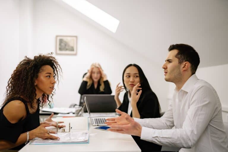 Vierköpfiges Team in angespannter Besprechung: zwei Personen gestikulieren im Gespräch, eine Kollegin im Hintergrund hält sich erschöpft den Kopf – Symbolbild für Überlastung und Kommunikationsdruck im Team.