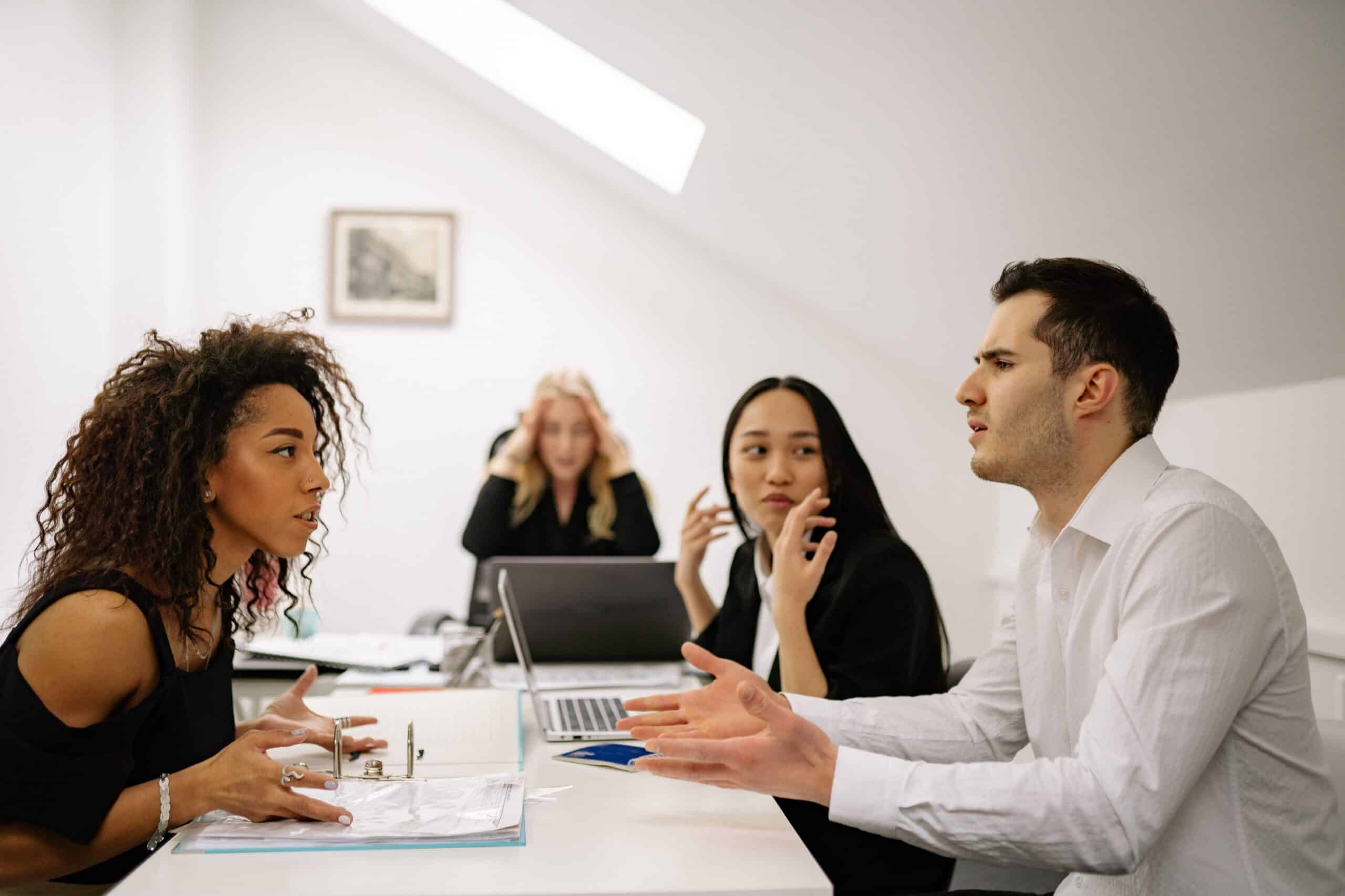 Vierköpfiges Team in angespannter Besprechung: zwei Personen gestikulieren im Gespräch, eine Kollegin im Hintergrund hält sich erschöpft den Kopf – Symbolbild für Überlastung und Kommunikationsdruck im Team.